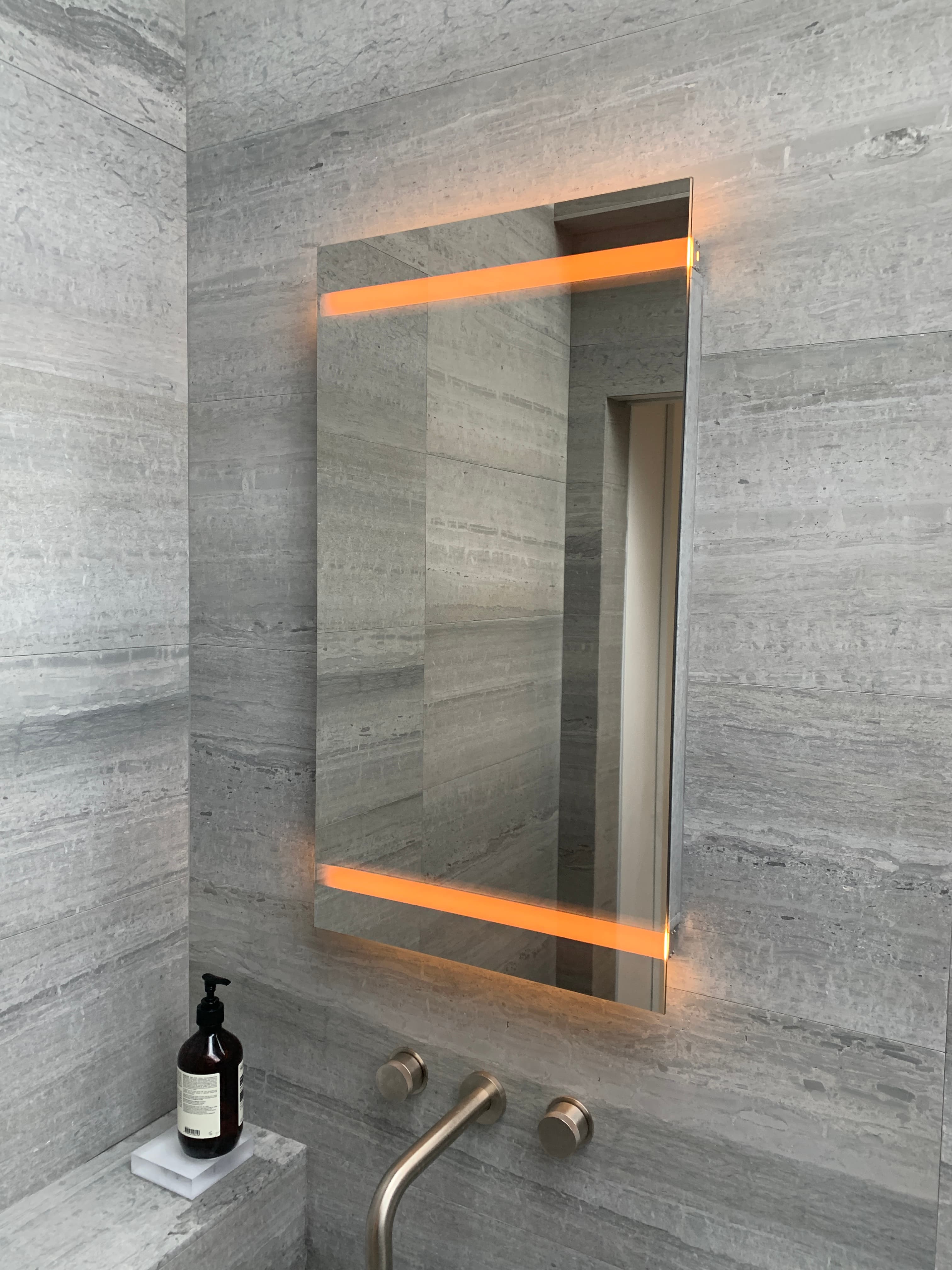 Backlit LED bathroom mirror mounted on grey stone tile above a vanity