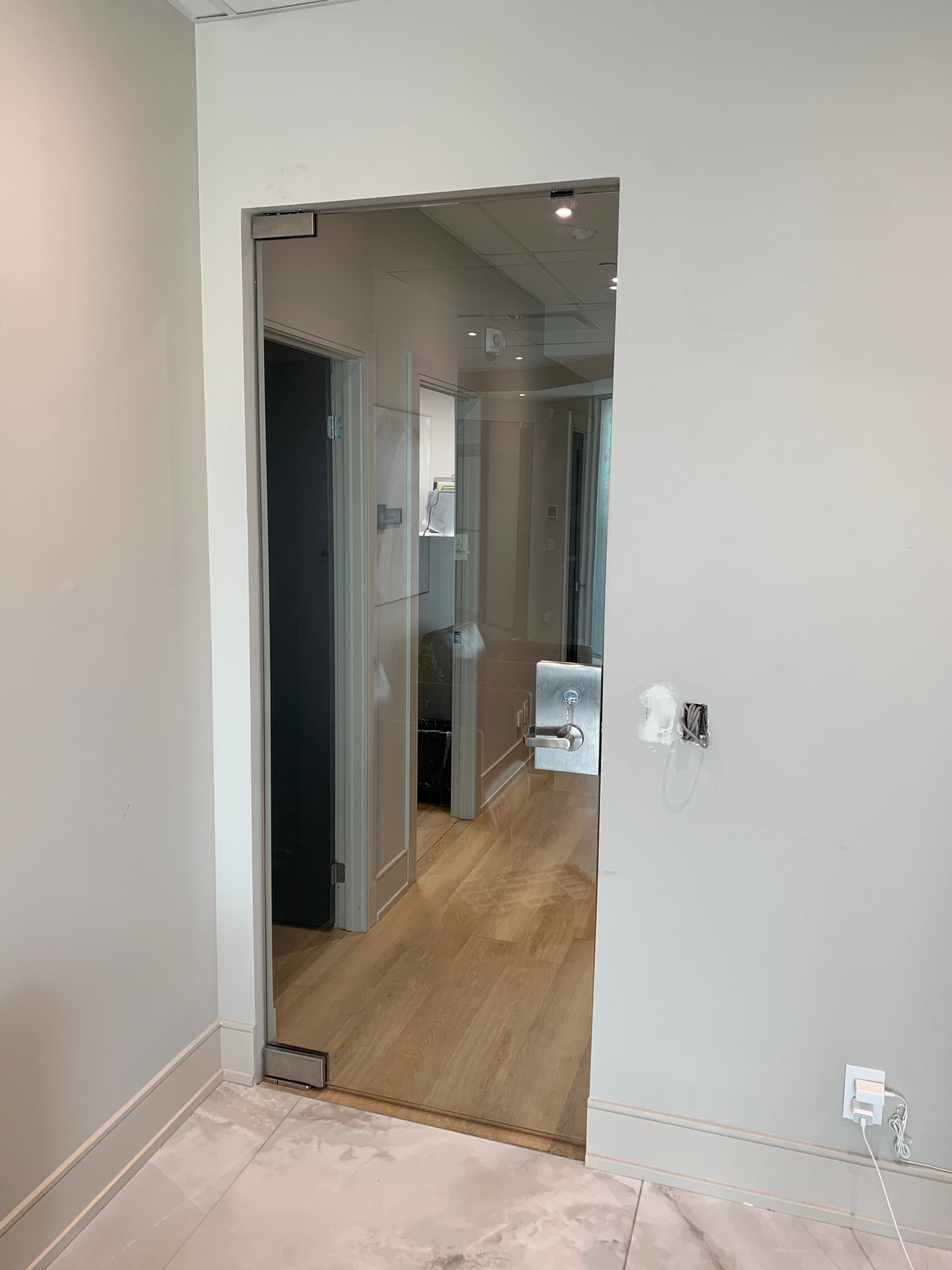 Frameless glass office door with polished pivot hinges and a keyed handle, opening onto a corridor.