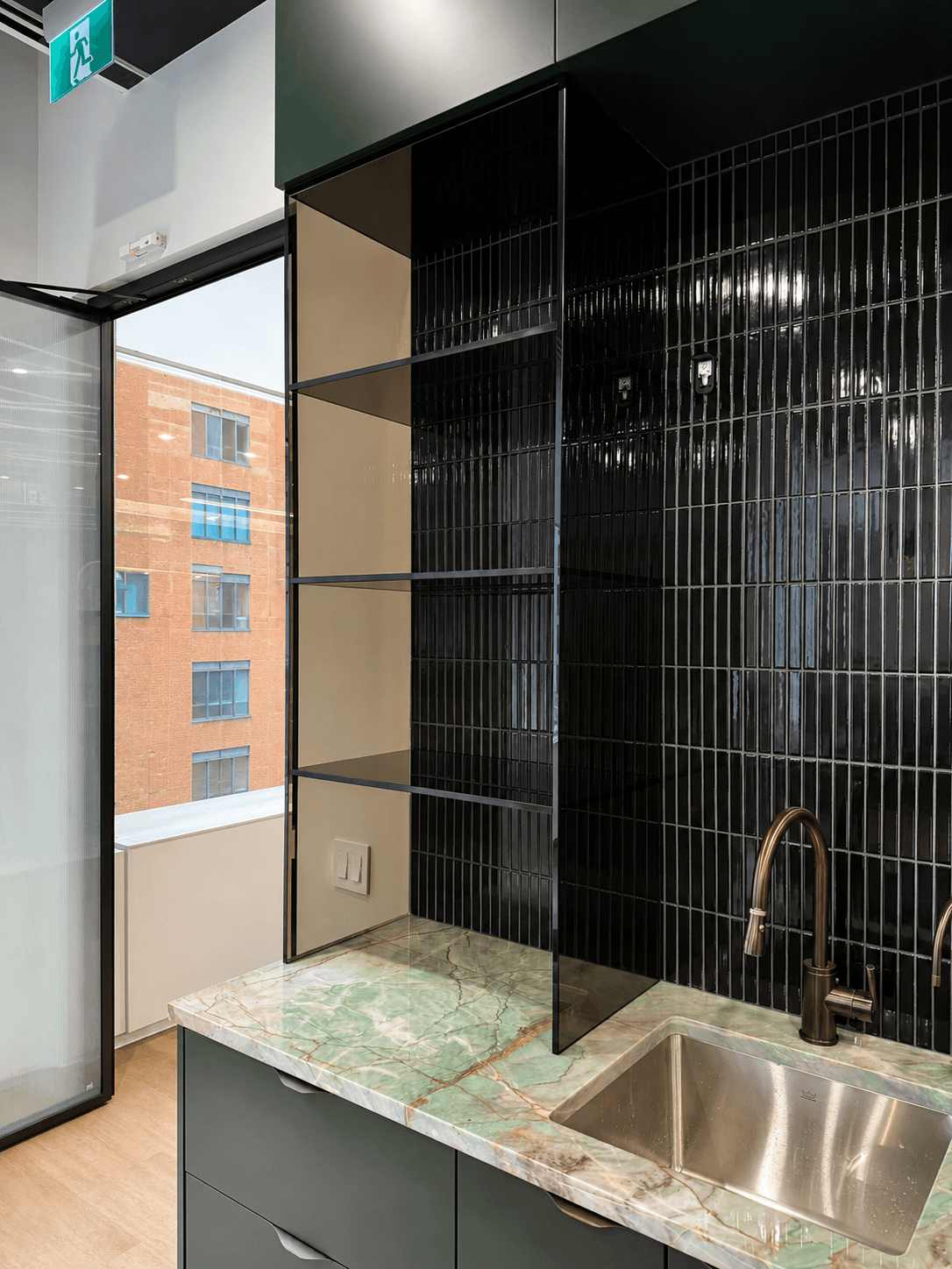 Office wet-bar nook with glass shelving against black tile, a green marble counter, and copper fixtures.