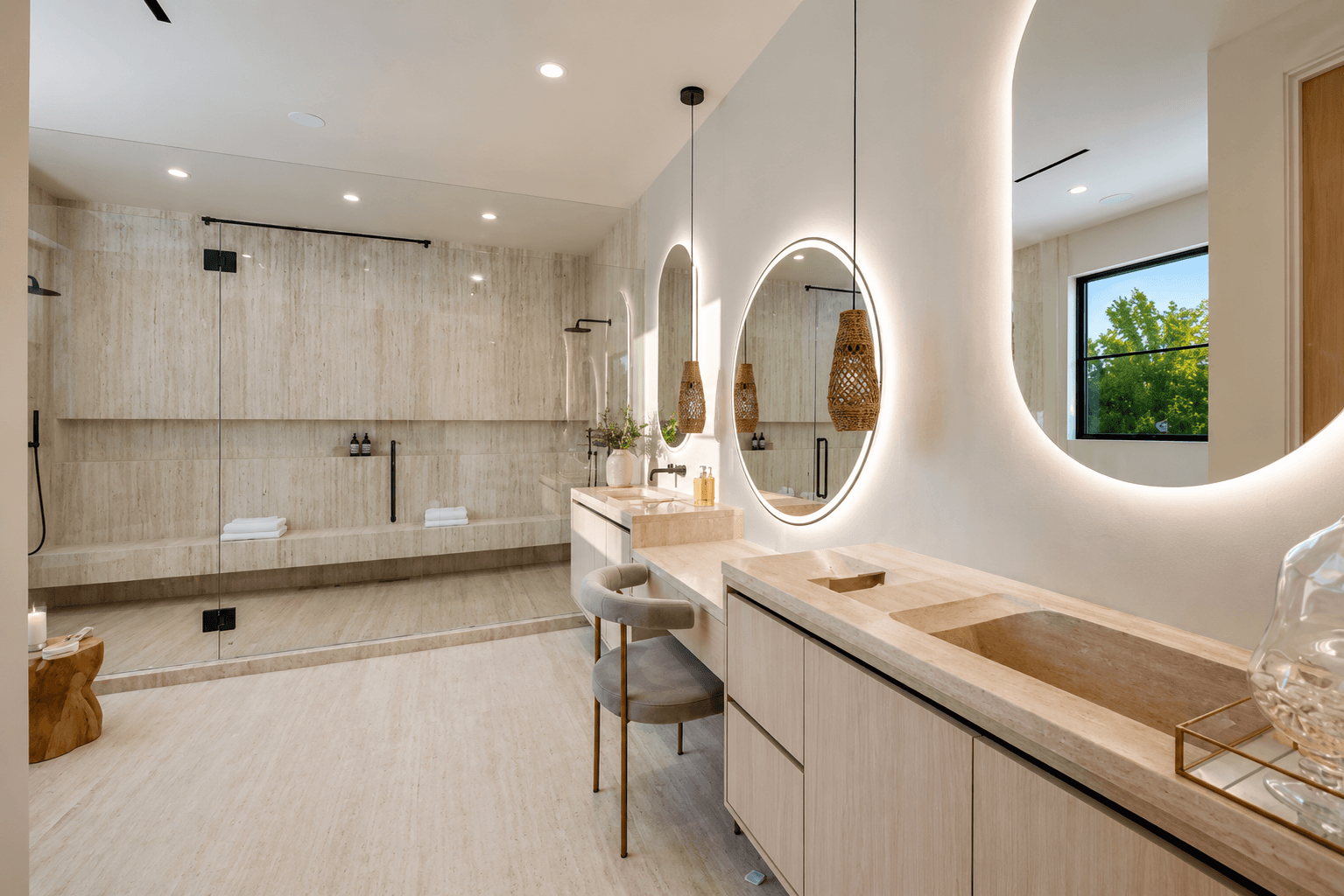 Primary bathroom with two round backlit LED mirrors above a travertine double vanity and glass shower.