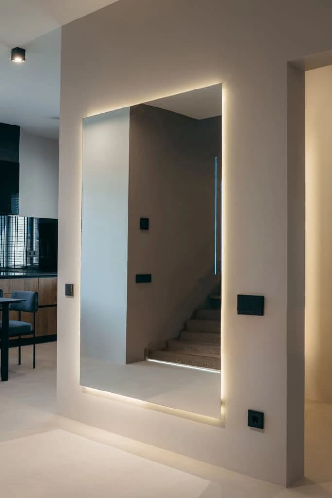 Large backlit frameless mirror set into a hallway wall, reflecting a staircase and kitchen.